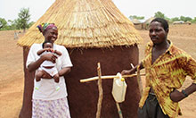 Woman, man, and baby stand outside a hut with a tippy tap