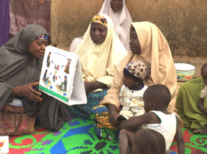 Photo of four women and two infants seated on a rug outside looking at a diagram.