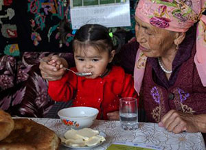 A grandmother helps her young granddaughter eat her food with a spoon.