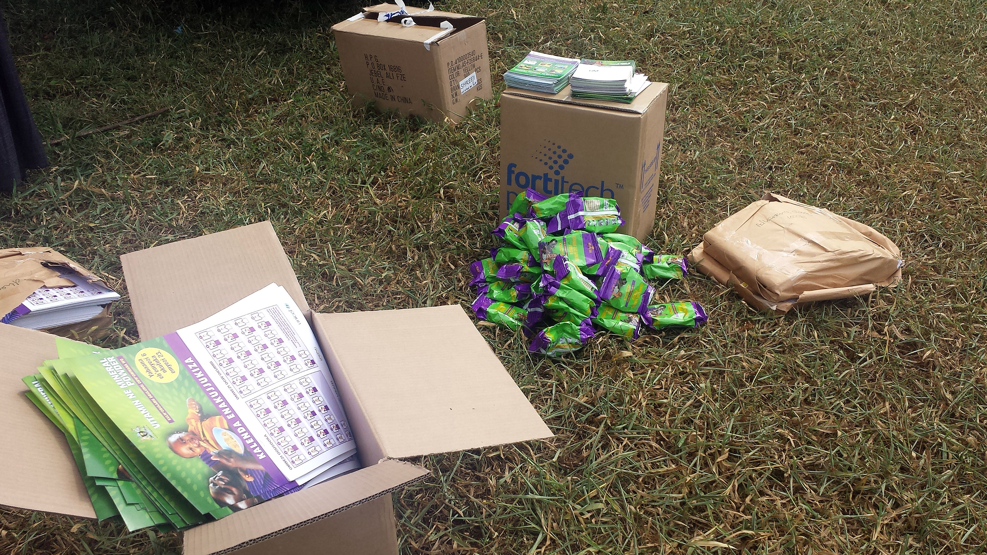 There are many materials to help with MNP distribution: adherence calendars (foreground), a pile of 60-day packaged supplies of MNPs (middle), boxes holding  MNP stickers and reminder cards (behind MNP pile). All of these materials, in addition to the M&E tools, are brought to the VHTs and health workers during distributions. After VHTs return to their villages, they distribute these materials to households, while health workers distribute these materials straight from the health facilities.