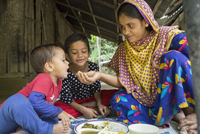 A Bangladeshi woman sits on a blanket with her two young children.