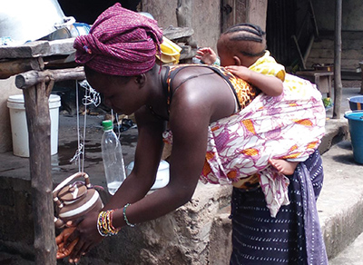  a mother in Sierra Leone bending down, carrying her infant child on her back as she uses a tippy-tap to wash her hands
