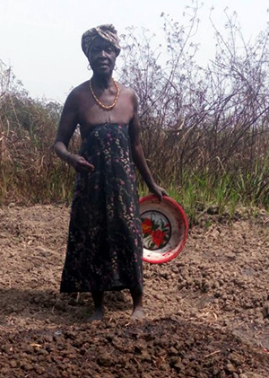 Photo of a woman standing on a bed of dirt, holding a bowl. 