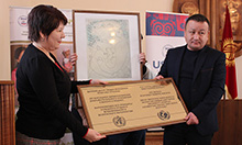 Photo of two people posing with a large plaque. USAID Health Project Management Specialist Aisha Zhorobekova presents the BFHI certification plaque to Abdulbaki Yrysbekov, Director of Karakul General Medicine Practice Center in Jalalabad oblast.