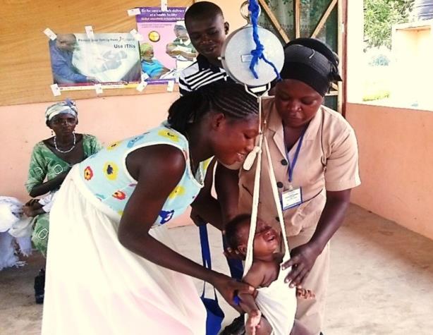 Photo of a community health worker weighing a baby at a child welfare clinic as the mother helps and the father and another family member watches in the background.