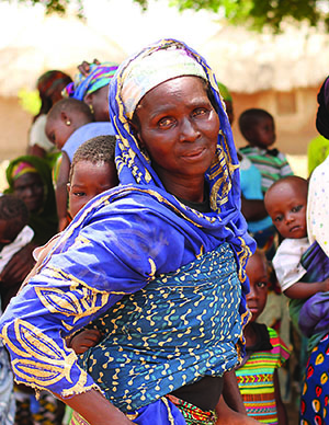 Photo of a mother standing in a crowd, smiling at the camera, carrying her child on her back.