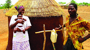 Photo of a man, a mother, and a child standing next to a tippy tap. Caption: Madam Ayaab of Yebonde community and her daughter are happy to get a follow-up visit from John, a Community Development Officer.