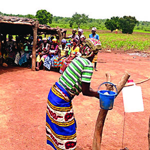 Photo of a woman demonstrating hwo to properly use a tippy tap. Caption: Tippy tap demonstrations in Dinyogu Community and Primary School - in Gushiegu District.