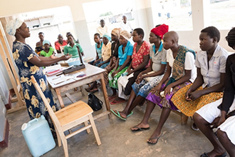 Photo of about a dozen or so pregnant women sitting in a room watching a woman give a demonstration/instruction. Caption: Pregnant women attend an antenatal care meeting in Amuria district. The NAWG included updated antenatal care guidelines in the anemia strategy