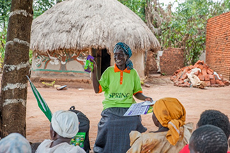Photo of a woman wearing a SPRING tee shirt giving a presentation outside to a group of mothers. Caption: "A VHT shows VMP sachets and reminder calendars to mothers in Namutumba district.