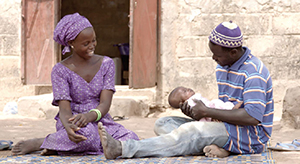 Photo of a man and woman sitting on a mat outdoors, with the man holding their infant child. 