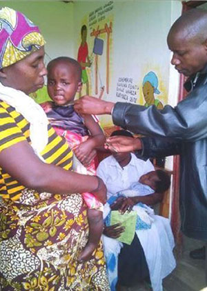 A health worker takes arm circumference and weight measurements on a 2-3 year old child being held by his mother. The child is making a distressed face.