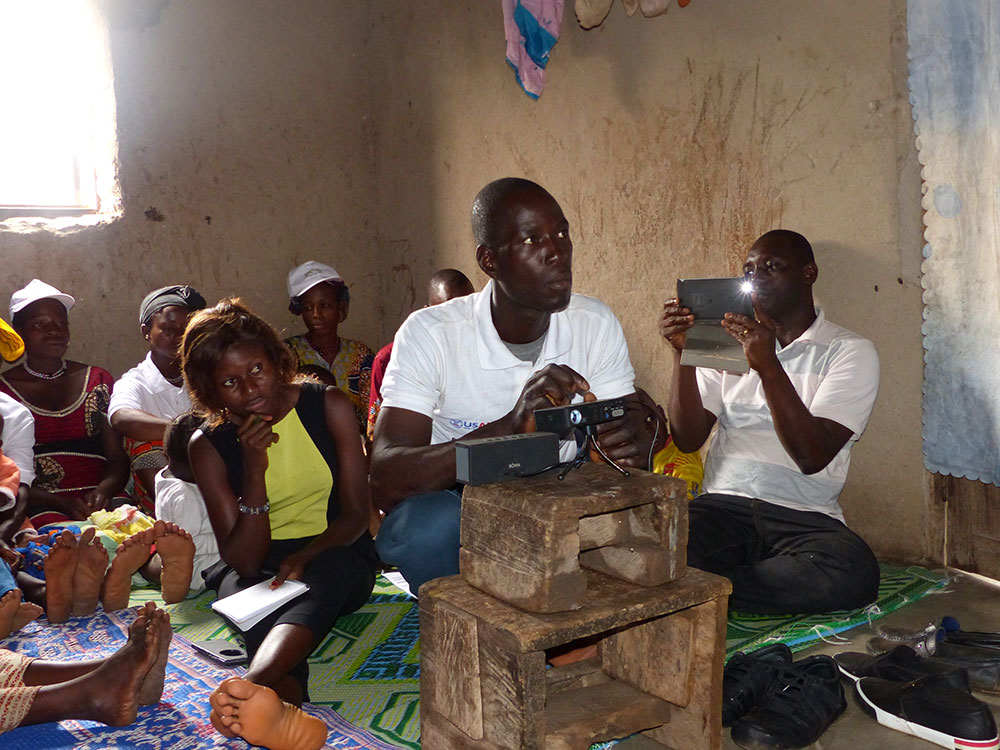 A SPRING/Guinea volunteer community worker sets-up a Pico projector to show the community video. SPRING/Guinea has developed community videos featuring exclusive breastfeeding practices, child nutrition, dietary diversity, handwashing with soap, benefits of consuming legumes, and processing potato leaves for off-season consumption.