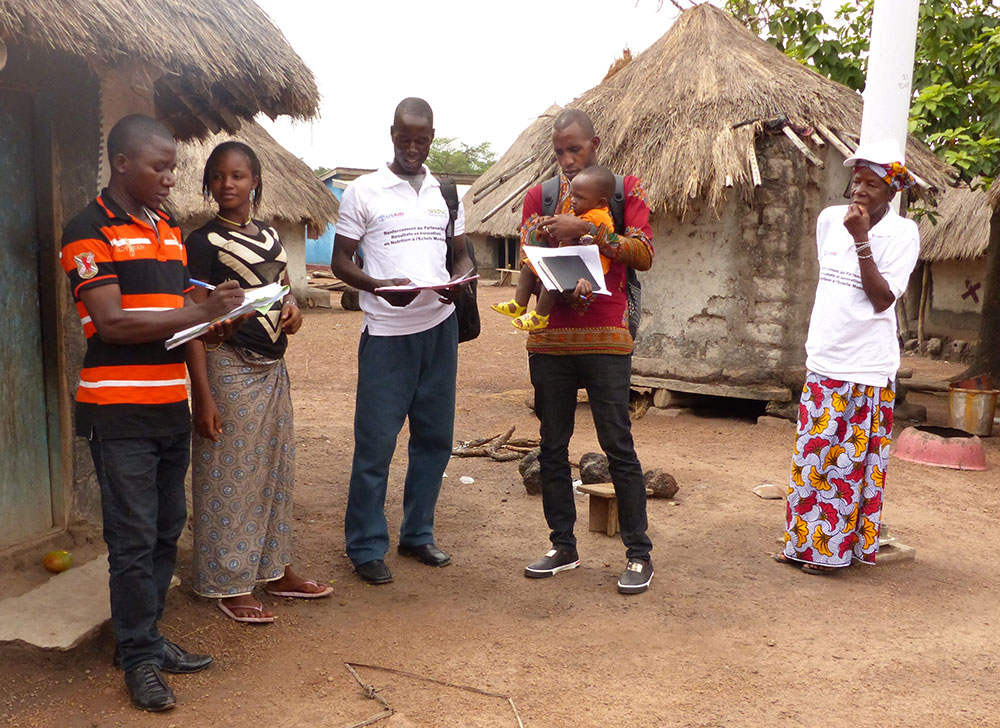 A SPRING/Guinea community volunteer in Bieindou demonstrates a home visit to the Sierra Leone visitors. Home visits reinforce messages promoted during the community video and assesses the family’s understanding of these key messages.