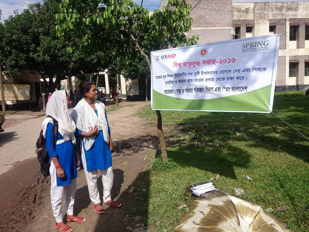 Two school girls stand next to a roadside banner in Abhaynagar upazila