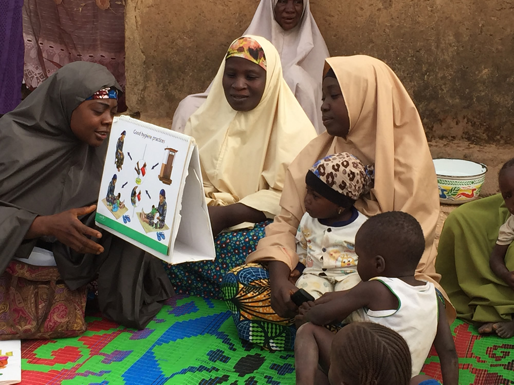 Young children accompanied their mothers while they listened to Mrs. Maryam Adams, a C-IYCF Community Volunteer in Kasuwan Magani ward in Kajuru LGA, discuss complementary feeding. C-IYCF Community Volunteers were observed to ask support group participants to explain the illustrations to ensure that they understood the messages. 