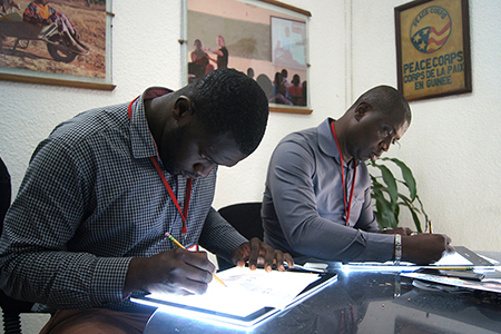 Photo of two men sitting at light tables, working on illustrations. Caption: "A light table helps the artist to easily see the lines from the printed photo through the back of the paper."