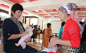 Photo credit: SPRING/Kyrgyz Republic. Two women sitting and talking in a large room with others doing the same. One woman holds cards about nutrition and is counselling the other women who is holding a realistic baby doll and practicing the proper way to hold an infant.