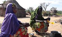 Two women crouch over a pot on a fire. One woman is stirring the moringa in the pot.
