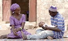 Photo of a man and woman sitting on a mat outdoors, with the man holding their infant child. 